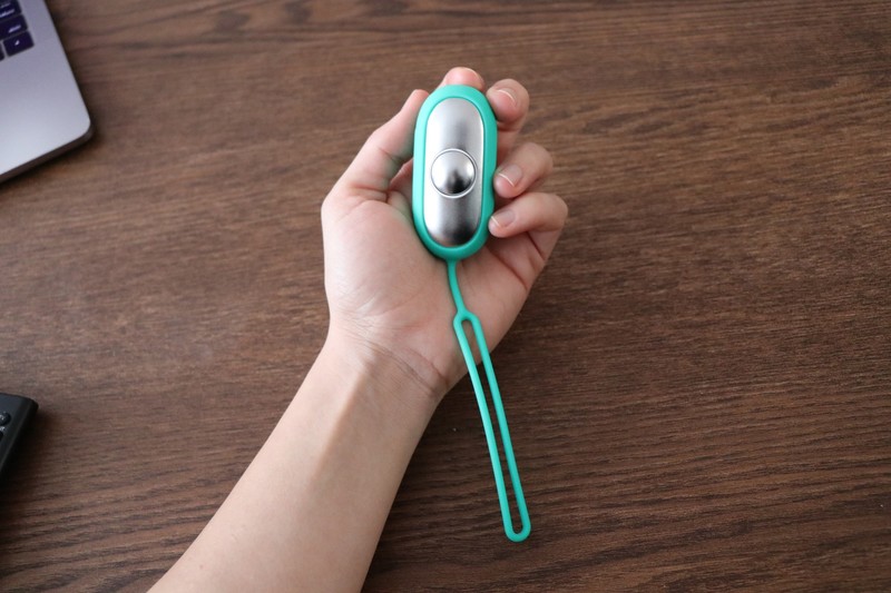 The Chill Pill calm device in teal, gripped in a hand at a desk showing the button and lanyard