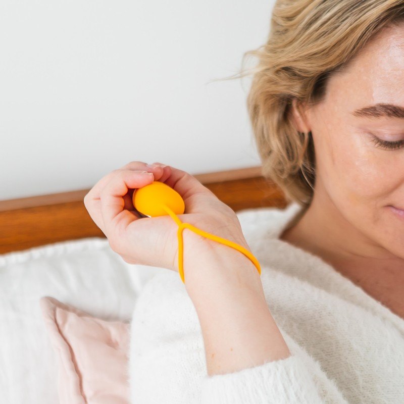 A person resting in bed holding the orange Chill Pill calm device by its wrist strap