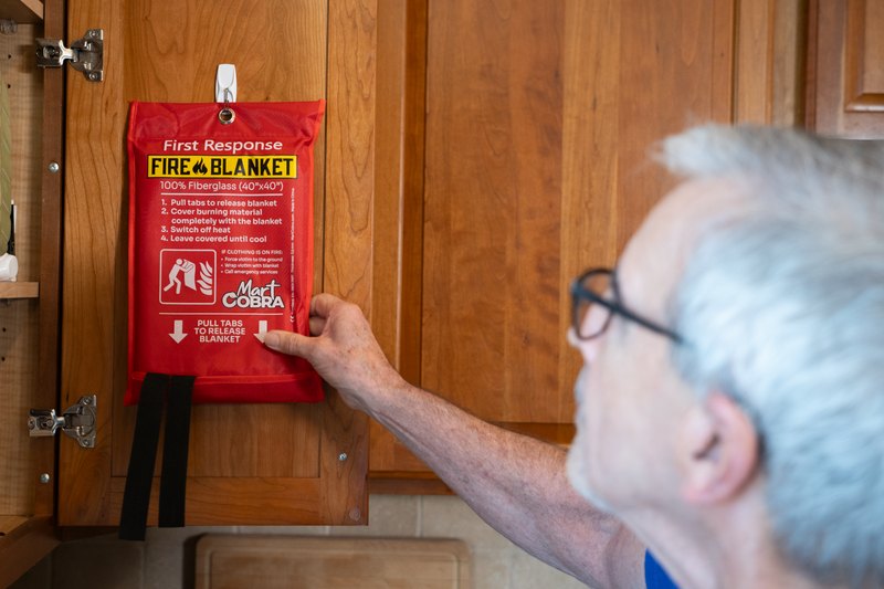 A hand pulling the release tabs on a wall-mounted Cobra Fire Blanket pouch in a kitchen