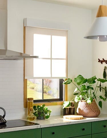 A Lutron Serena roller shade lowered over a kitchen window in a bright, plant-filled room