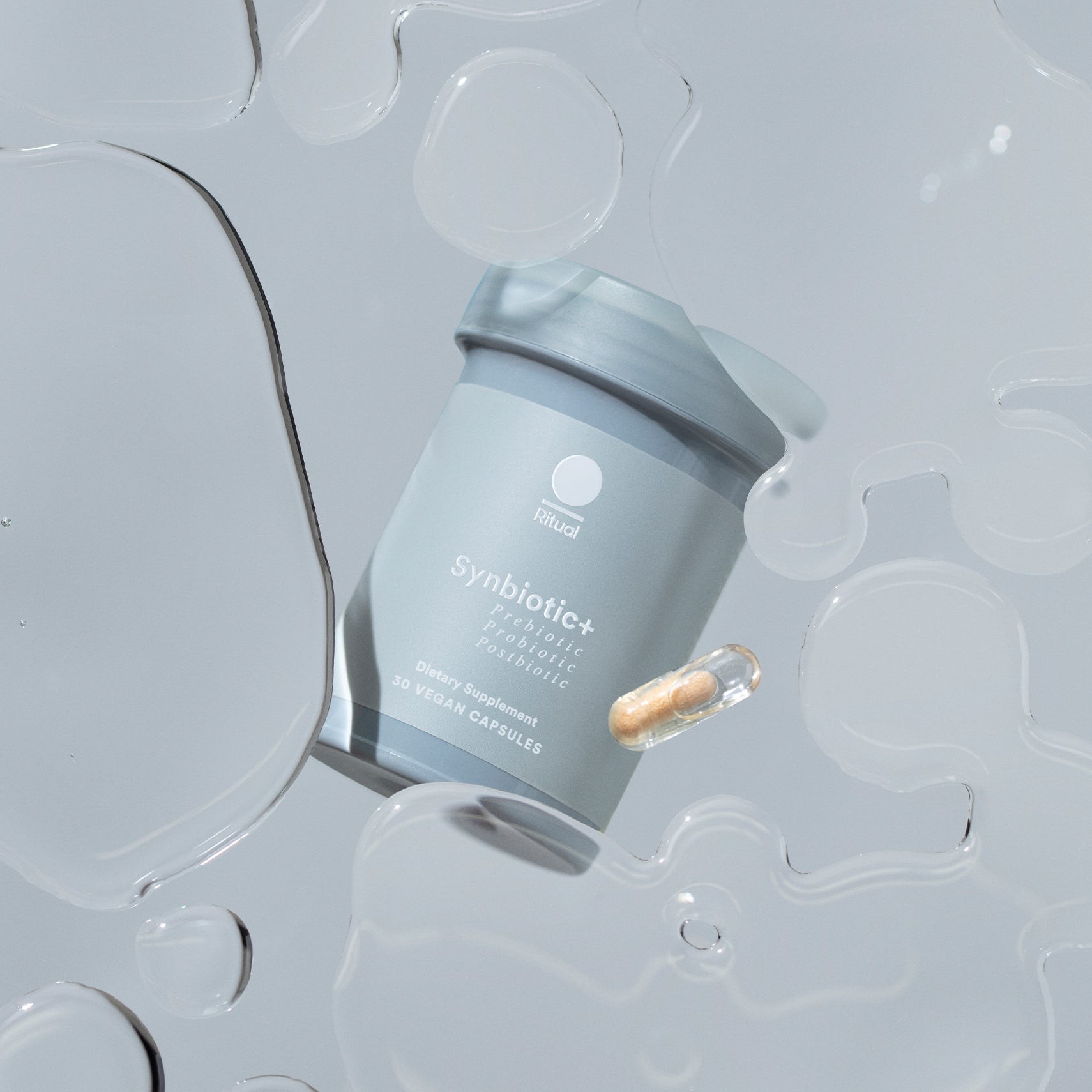 A grey Ritual Synbiotic+ capsule bottle with a single two-tone capsule on a water-droplet background