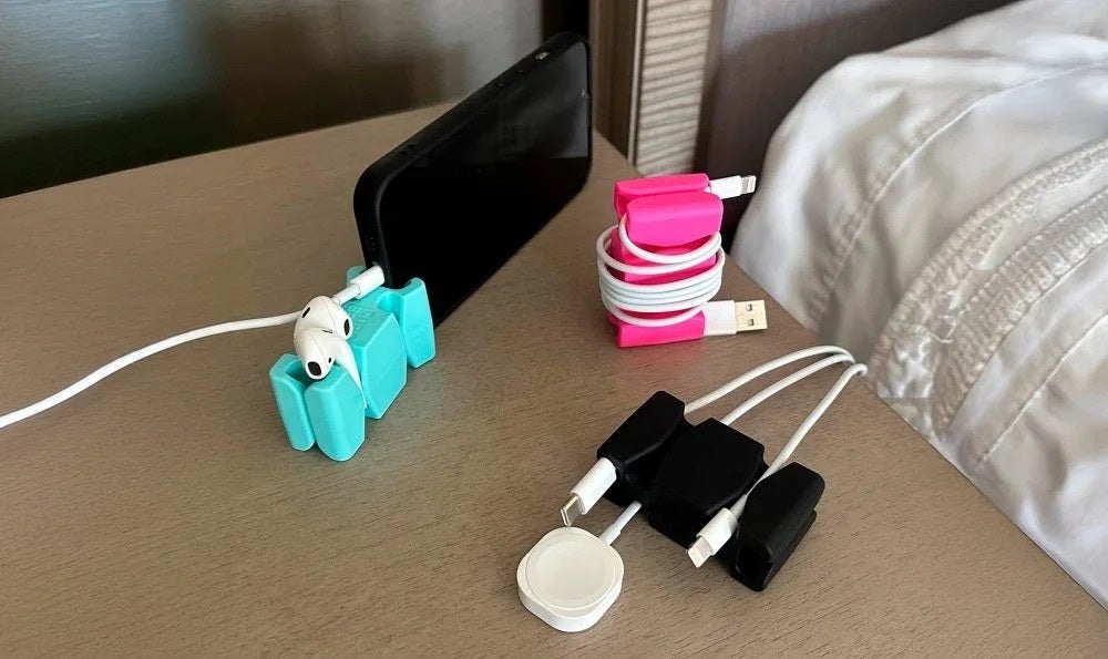 CordBrick weighted silicone cord holders on a nightstand, one holding a phone upright with earbuds, others keeping charging cables in place
