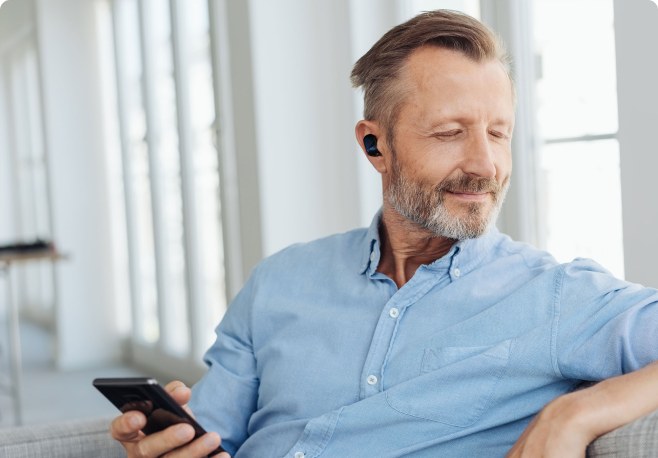 A man wearing a black Echo Labs earbud while relaxing on a couch and looking at his phone