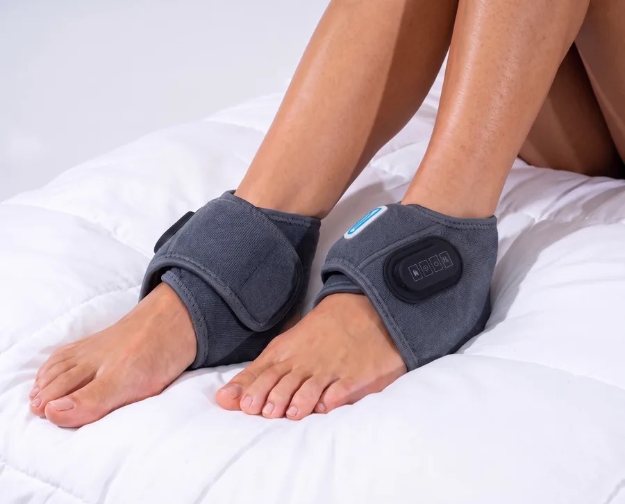 A pair of EMSense EMS foot massager wraps worn on both feet resting on a bed