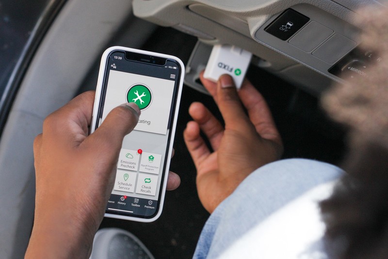 A driver tapping the FIXD app on a smartphone while holding the FIXD sensor near a car's diagnostic port