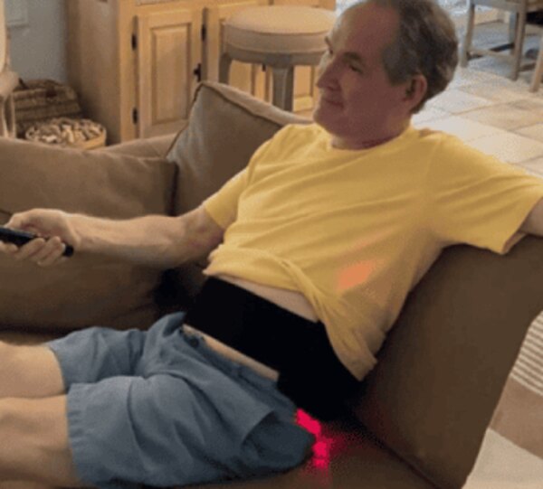 Person relaxing on a couch with the Glokore Red Light Therapy Wrap around the lower back, holding the remote