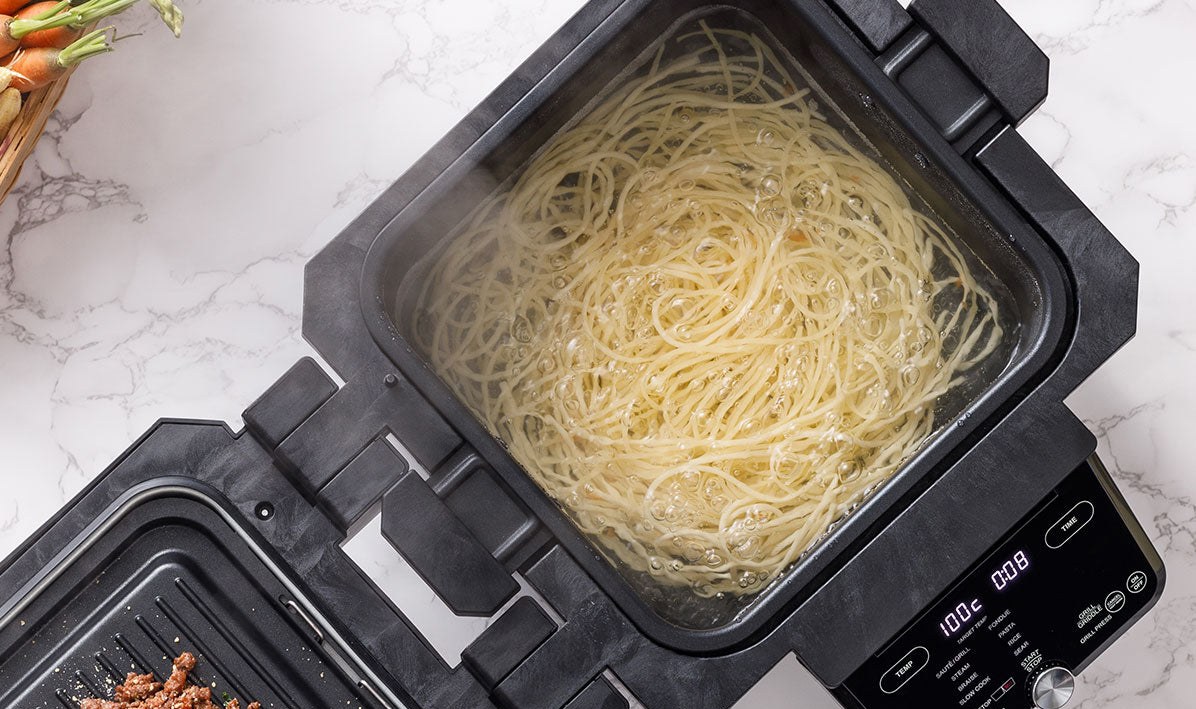 The Gourmax Pro cooking pasta in one open tray while bolognese sits on the grill plate, showing its dual cooking surface