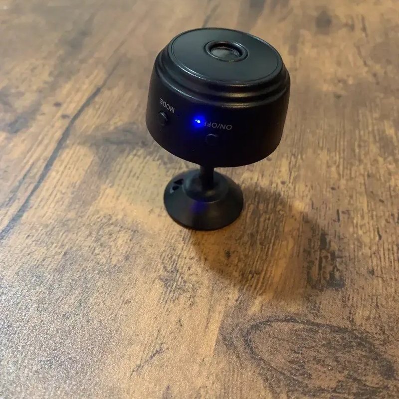 Home Defender Cam security camera sitting on a wooden table with its blue status light on