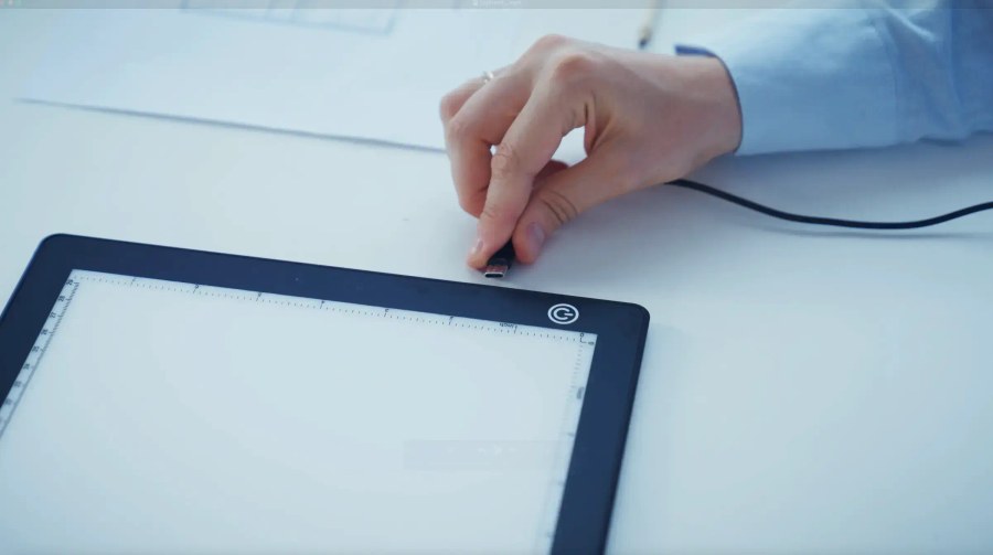 Lite Sketch Lightpad on a desk with a hand plugging in the USB-C cable, showing the ruler edge and power button