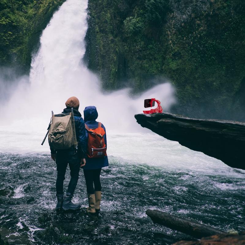 Two travelers filming themselves with a phone mounted on a red Tenikle on a rock ledge by a waterfall