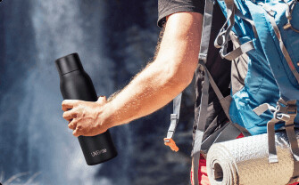 A person holding the black UVBrite Smart Bottle outdoors near a waterfall with a hiking pack