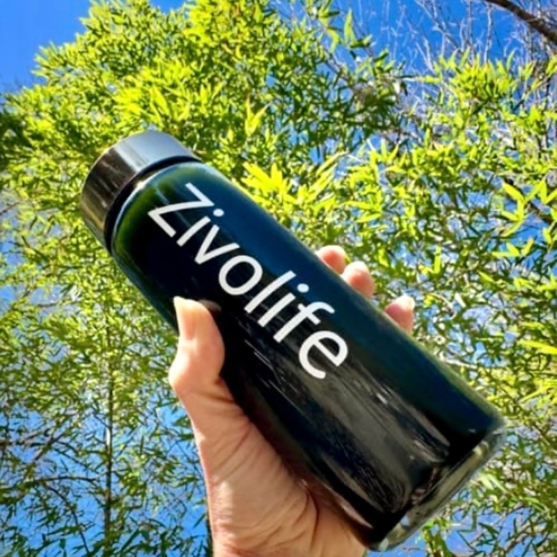 Hand holding a ZivoLife drink bottle outdoors against green trees and blue sky